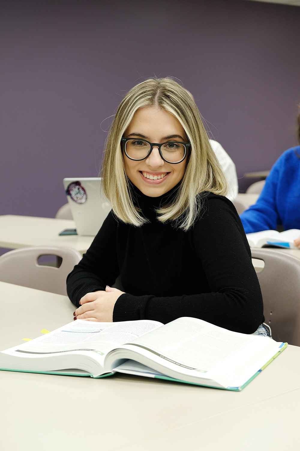 A woman sitting at a desk looks up from her textbook to smile at the camera.
