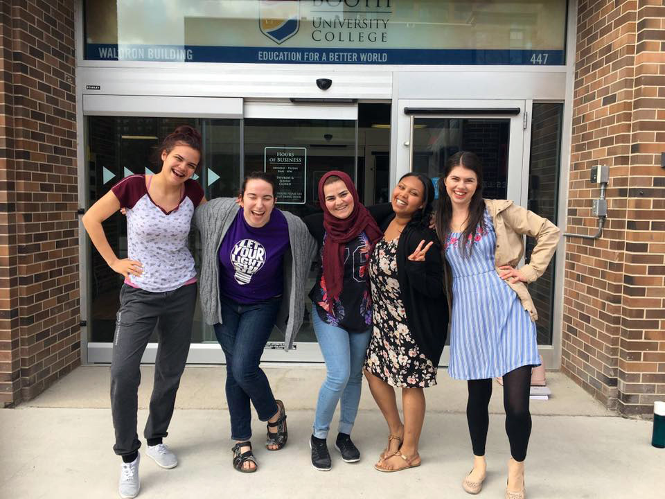 Five female students stand with arms around each other in front of the school.