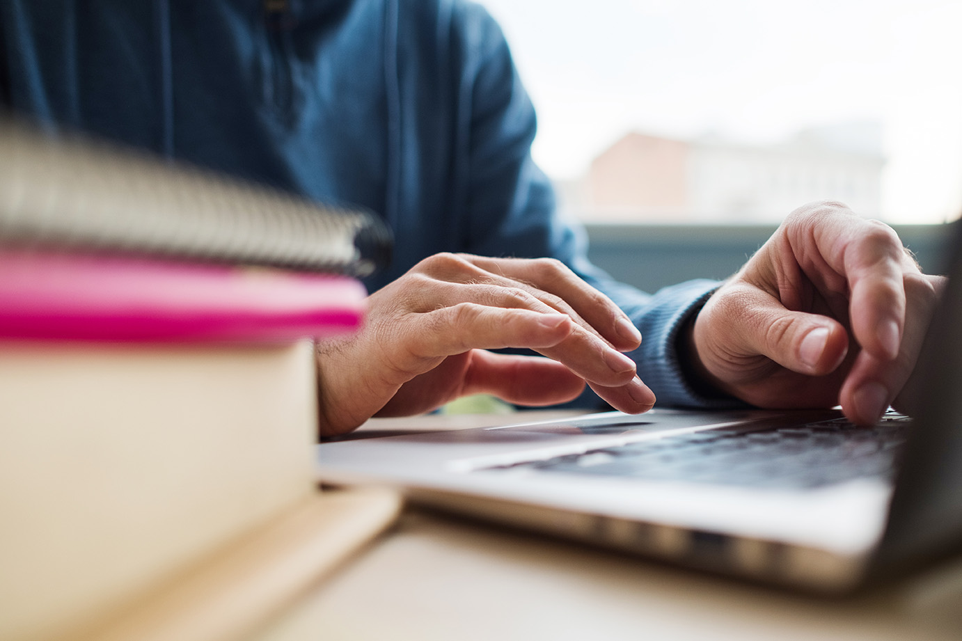 Hands typing on a laptop computer beside a stack of textbooks.