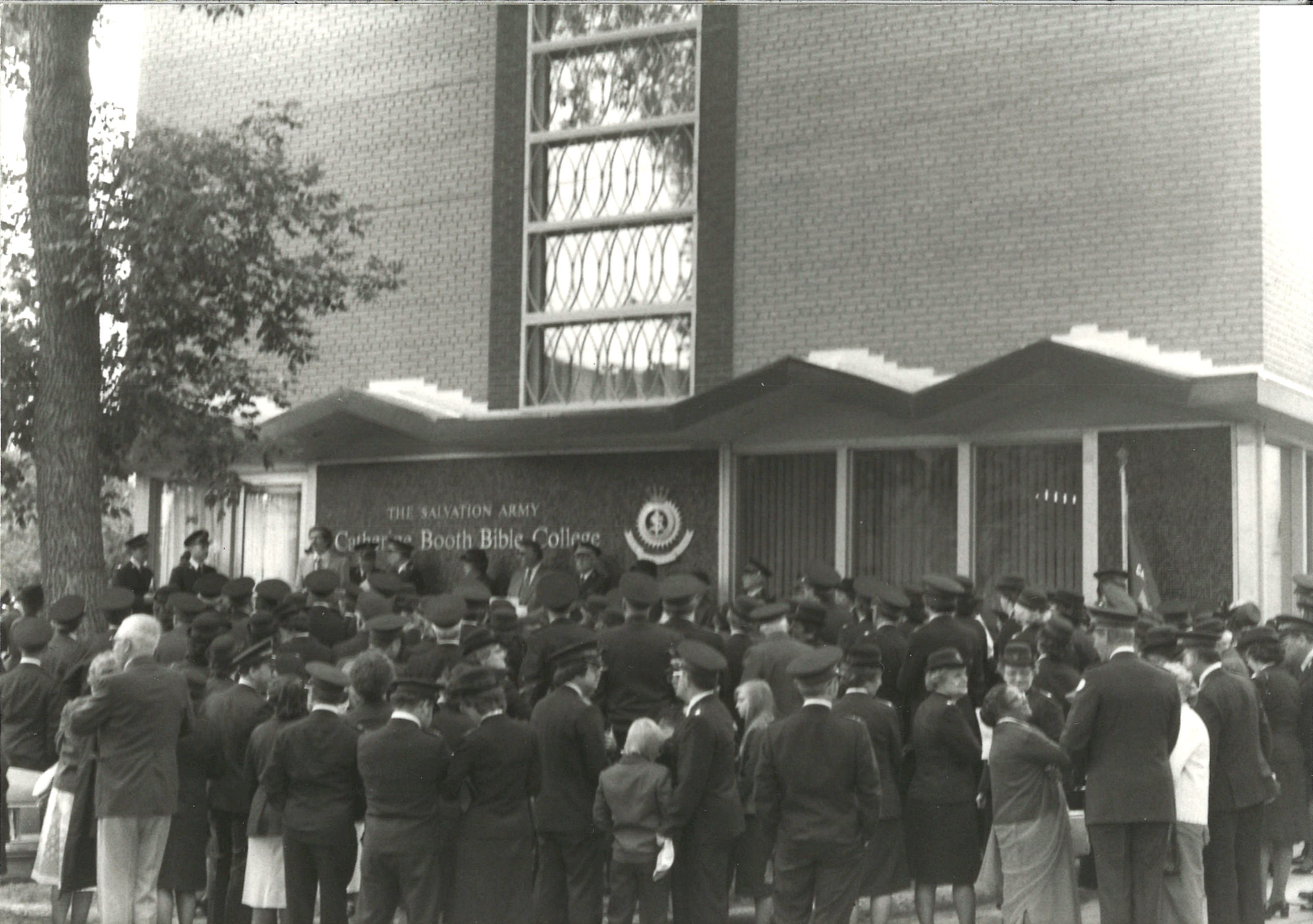 In black and white, people gather outside the front entrance for a ceremony at the first property.