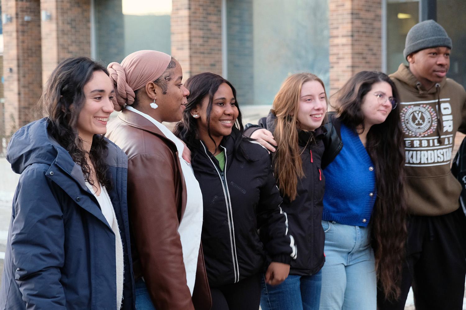 Students stand with arms around each other outside the campus entrance.