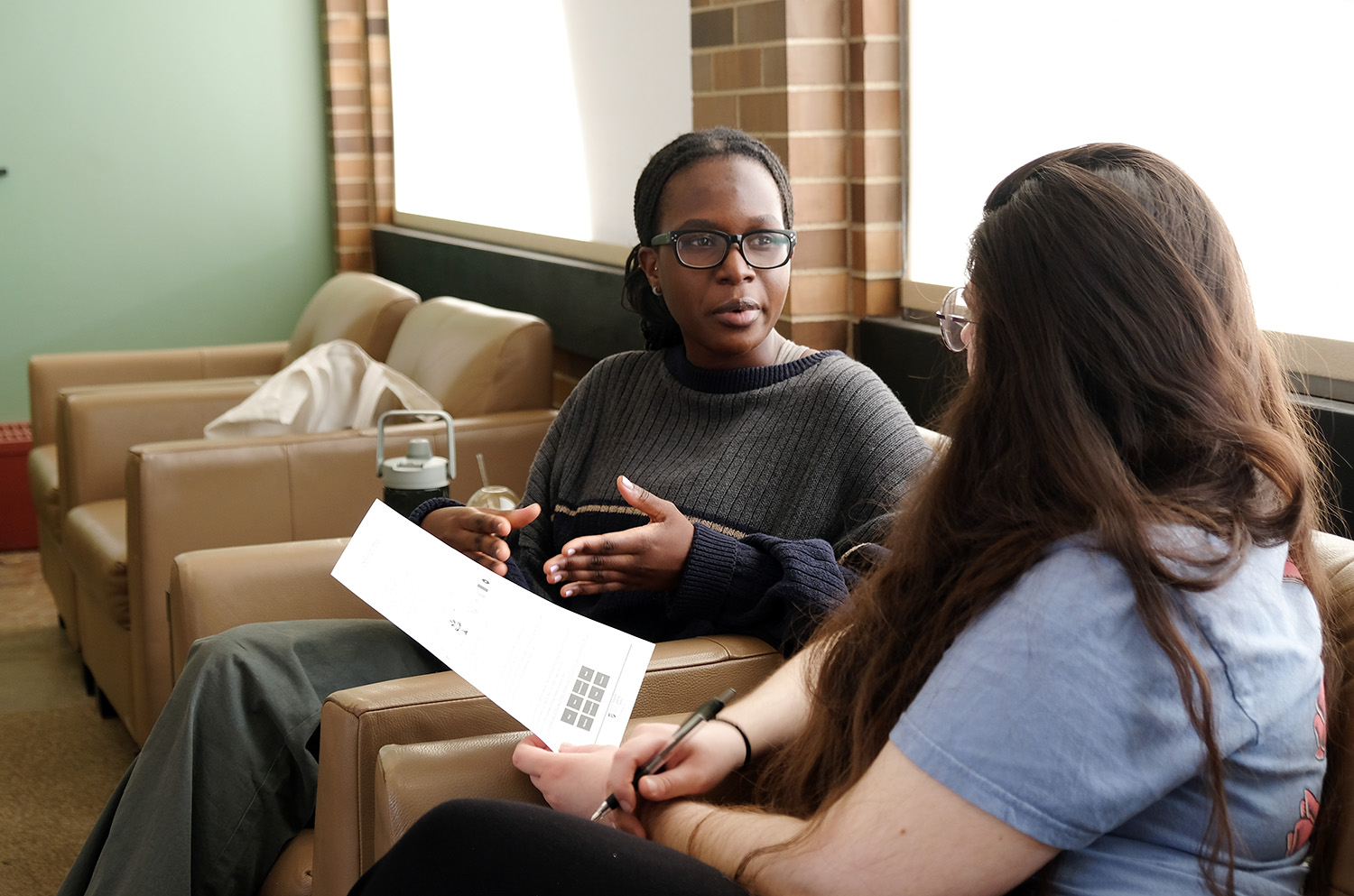 Two students sitting in comfortable lounge chairs in front of a window, holding papers and talking.