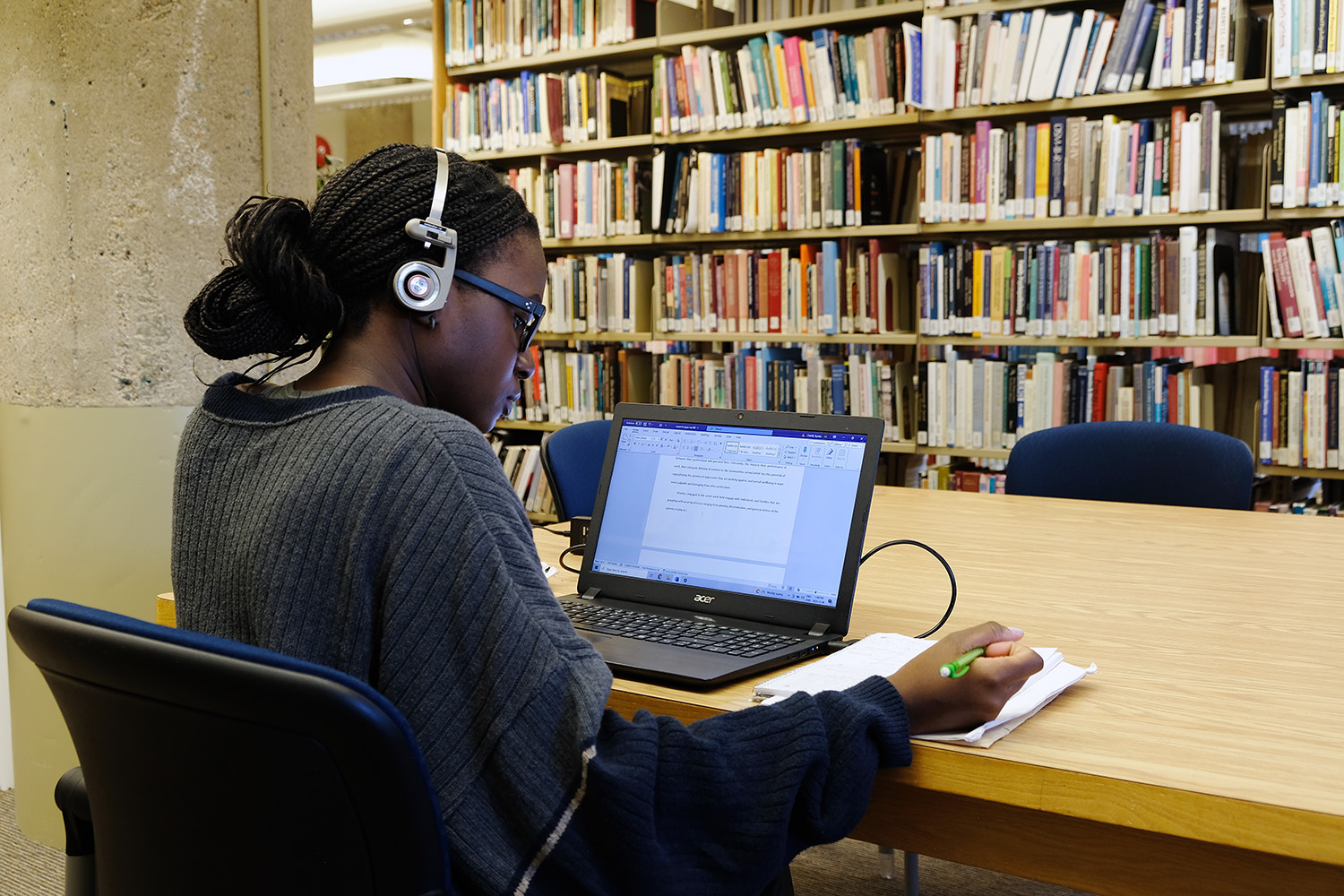 Female student working in a library writing in a notebook with her laptop open on the desk