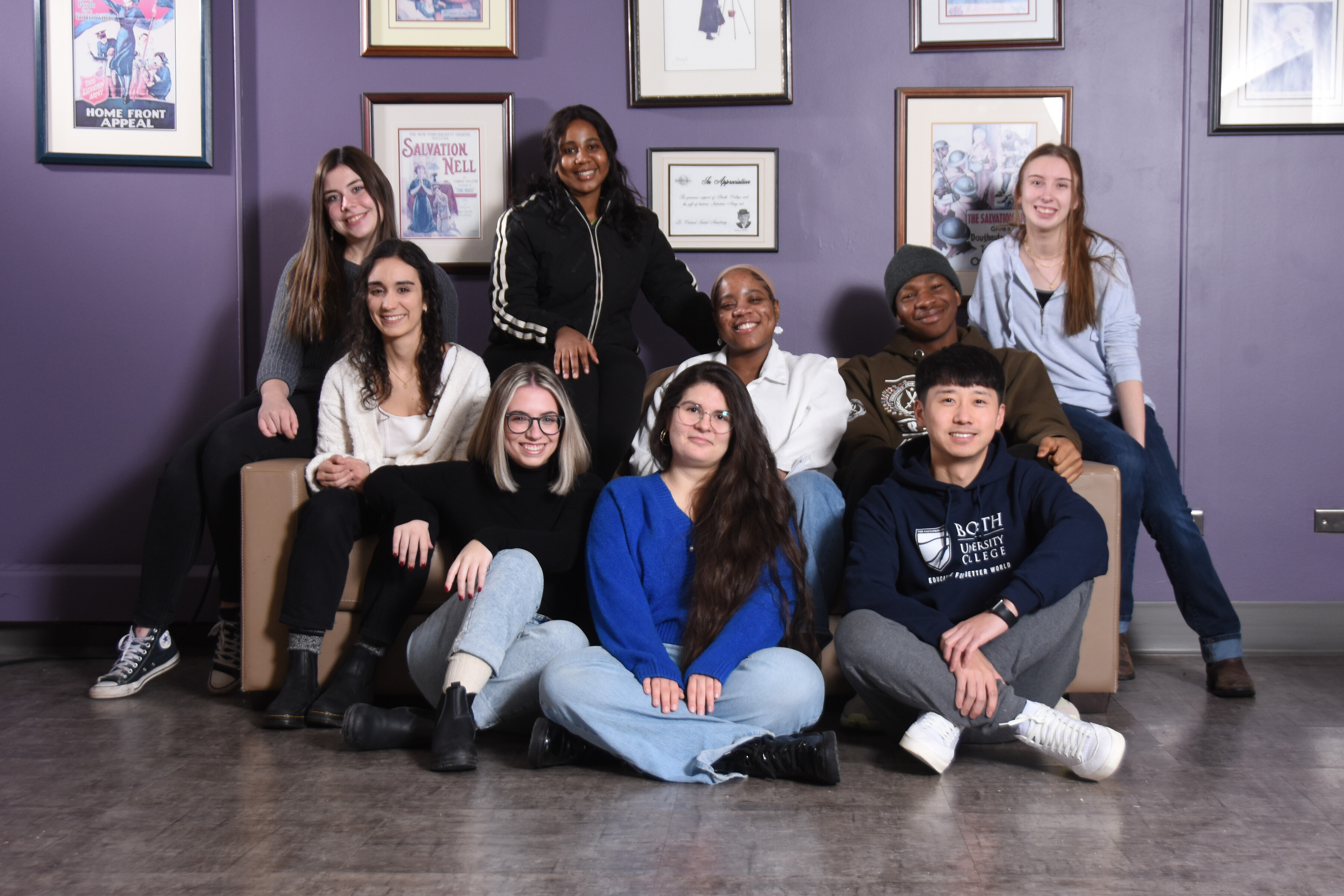 Nine students pose on a sofa, some sitting on the floor in front and some across the back.