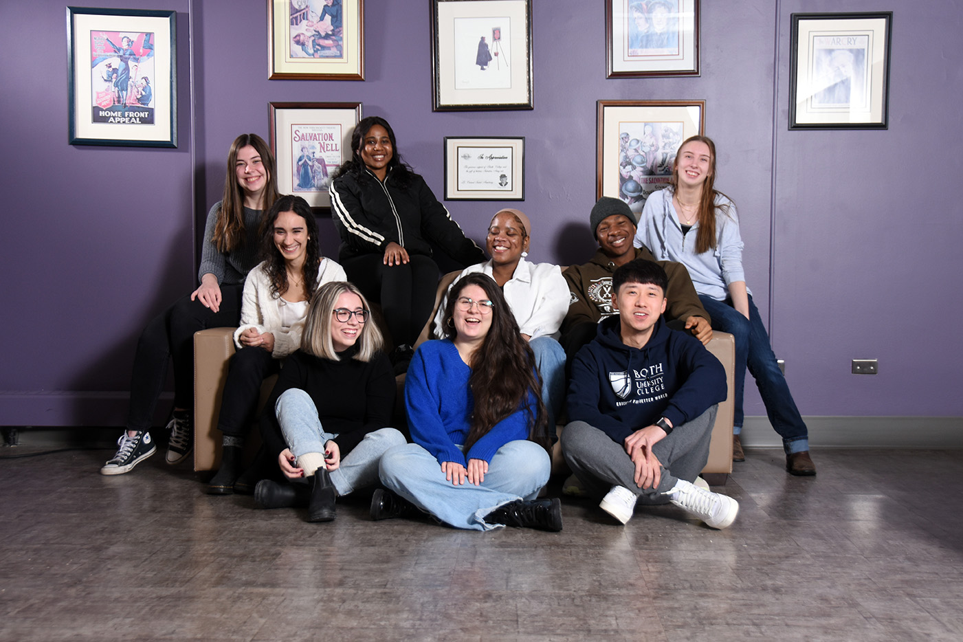 Nine students pose on a sofa, some sitting on the floor in front and some across the back.