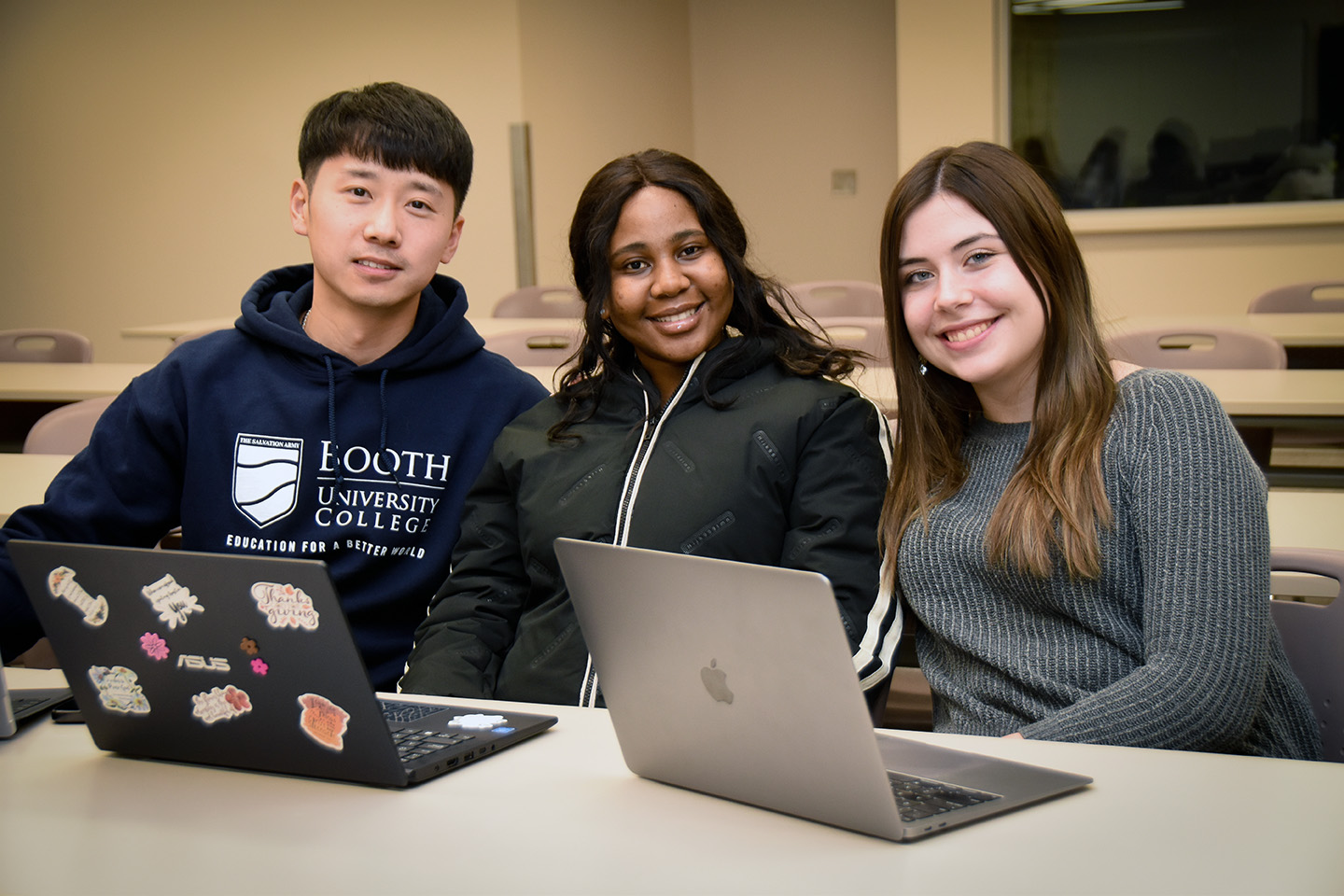 Three students sitting at a desk in a classroom. Each of them has a laptop and is smiling at the camera.