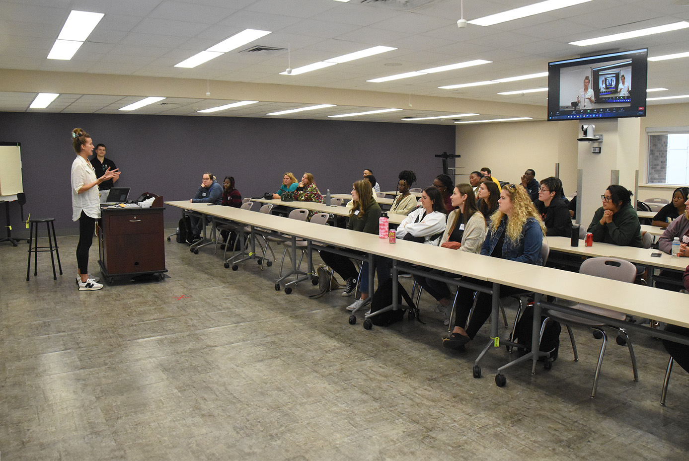 An instructor stands at the front of a large classroom speaking to students at long desks.