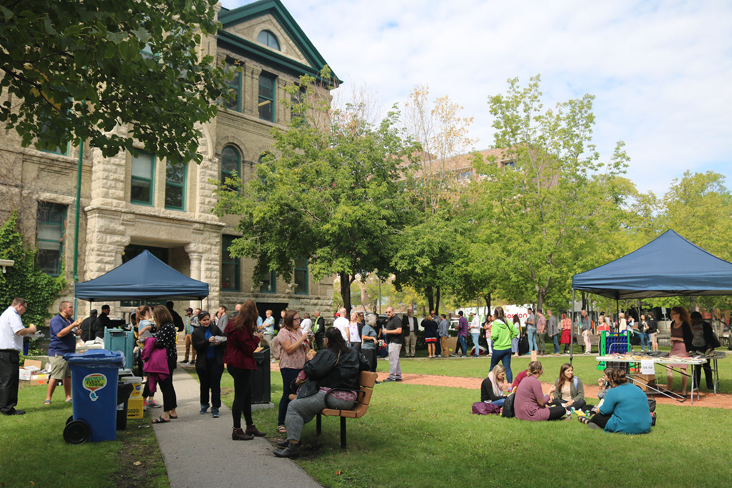 People gathered in green space outside the campus. Some people are sitting in small groups on the grass and others are standing and talking.