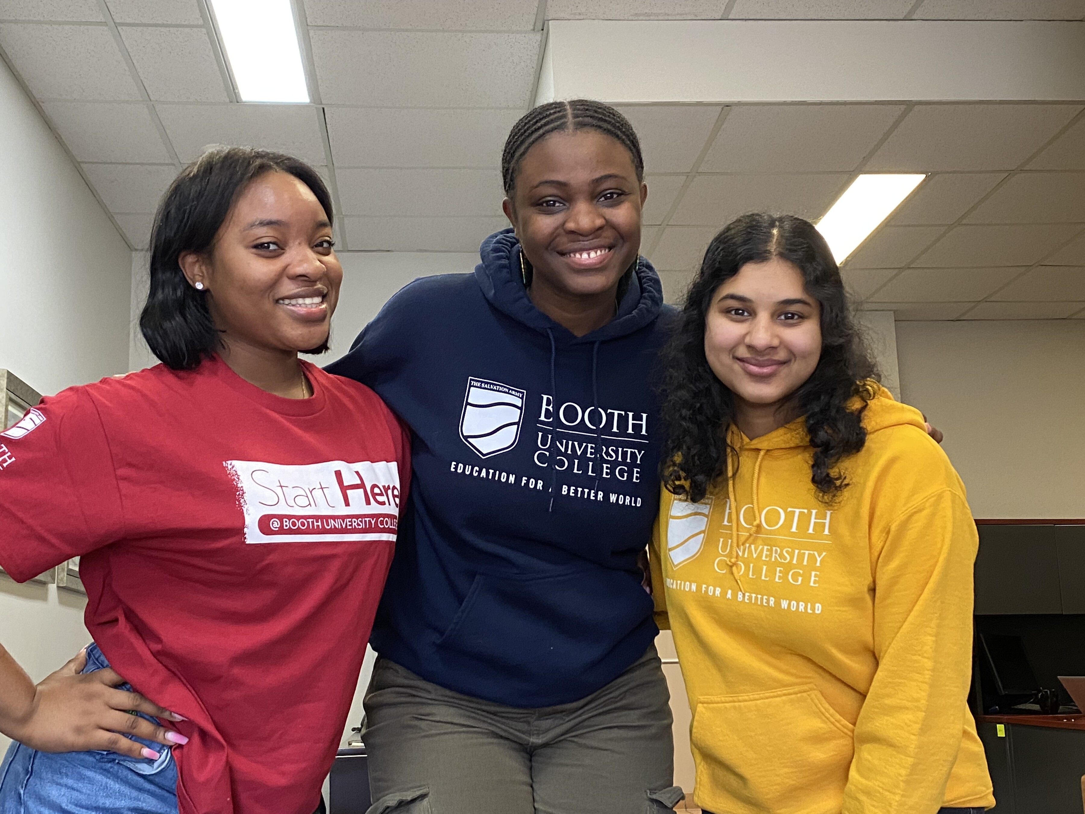 Three students are wearing red, blue, or yellow shirts with the Booth University College logo across the fronts.